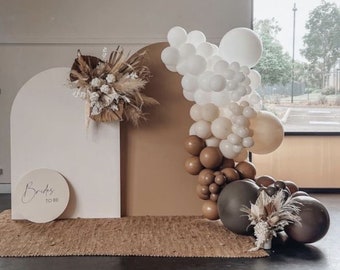 May include: A wedding backdrop featuring a cream-colored arch and a tan wall, adorned with a balloon garland in white, tan, and brown. Dried floral arrangements and a circular sign that reads "Brides to be" complete the decor.