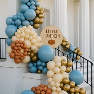 May include: A festive balloon arch in shades of blue, cream, gold, and brown decorates a white exterior. A wooden sign with the text "Little Pumpkin" and a pumpkin graphic is featured. The balloons are arranged on steps.