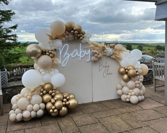May include: A beige backdrop with a neon sign that says "Baby" and a handwritten sign that says "Baby Cann". The backdrop is decorated with a garland of white, beige, and gold balloons.