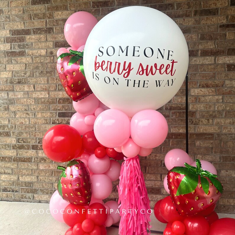 Peut inclure: Un ballon blanc avec le texte "Someone berry sweet is on the way" en lettres rouges et noires. Le ballon est entour&eacute; de ballons roses et rouges, certains avec des motifs de fraises. Il y a aussi un pompon rose suspendu au bas du ballon.