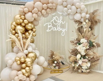 May include: A gold and cream balloon arch with pampas grass and flowers, decorated with the text "Oh Baby!" in neon lights.