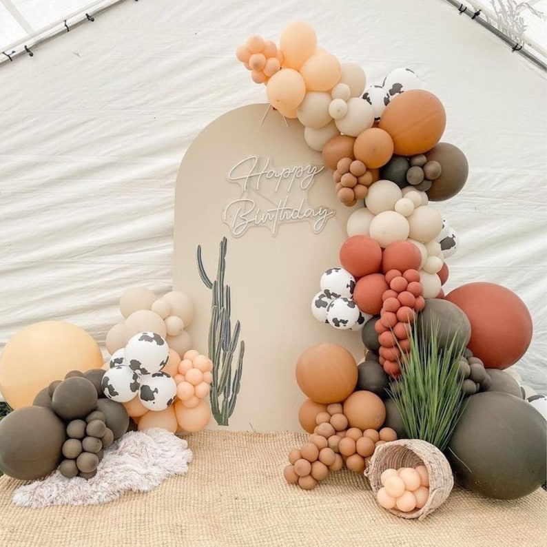 Pu&ograve; includere: Un arco beige con le parole "Happy Birthday" in luci al neon, decorato con una ghirlanda di palloncini marroni, beige e bianchi. I palloncini sono disposti a cascata e includono disegni di mucca. Ci sono anche palloncini marroni, beige e bianchi sul pavimento davanti all'arco.
