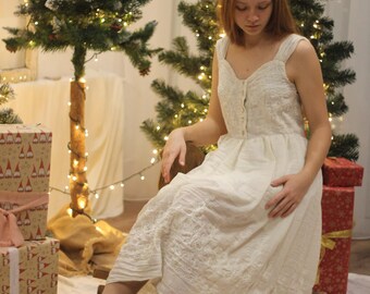 May include: A young woman wearing a white dress with embroidered details sits in front of a Christmas tree with white lights. She is holding a gift box in her lap.