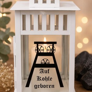 Könnte beinhalten: Eine weiße Metalllaterne mit einer Glasscheibe, die eine schwarze Grafik eines Förderturms und den schwarzen Schriftzug "Auf Kohle geboren" zeigt. Im Inneren der Laterne befindet sich eine weiße Stumpenkerze mit einer sichtbaren Flamme.