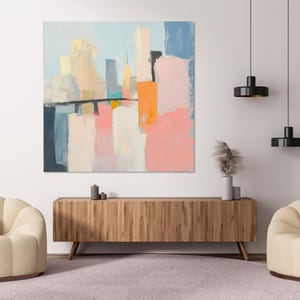 Könnte beinhalten: Abstrakte Malerei eines Stadtbildes in Blautönen, Rosa, Gelb und Weiß. Das Gemälde hängt an einer weißen Wand über einem Holzschrank.