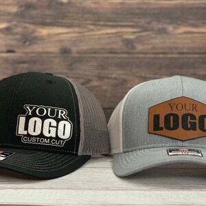 May include: Two black and gray trucker hats with white mesh back panels. One hat has a white embroidered logo that says "YOUR LOGO CUSTOM CUT". The other hat has a brown leather patch with the text "YOUR LOGO" in black.