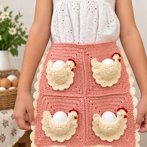 May include: A pink crochet apron featuring four integrated pockets shaped like cream-colored chickens, each holding a white egg. The apron has a scalloped cream-colored border and is constructed from four square panels joined together.