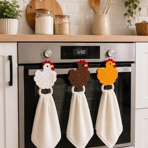 Könnte beinhalten: Drei gehäkelte Hühner-Handtuchhalter in Weiß, Braun und Gelb, jeweils mit rotem Kamm und Kehllappen. Jedes Huhn ist an einem hellen Holzring befestigt, der ein schlichtes, weißes, rechteckiges Küchentuch hält und am Ofengriff hängt.