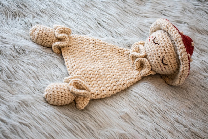 May include: A crocheted doll with a red and white hat, wearing a beige crocheted outfit. The doll is lying on a fluffy white surface.