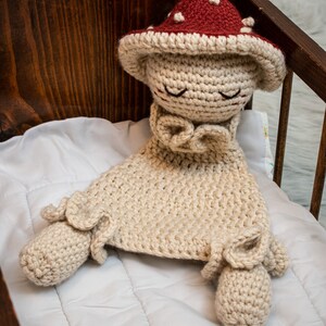 May include: A crocheted mushroom-shaped baby toy with a red and white hat, lying in a wooden crib with a white quilted mattress.