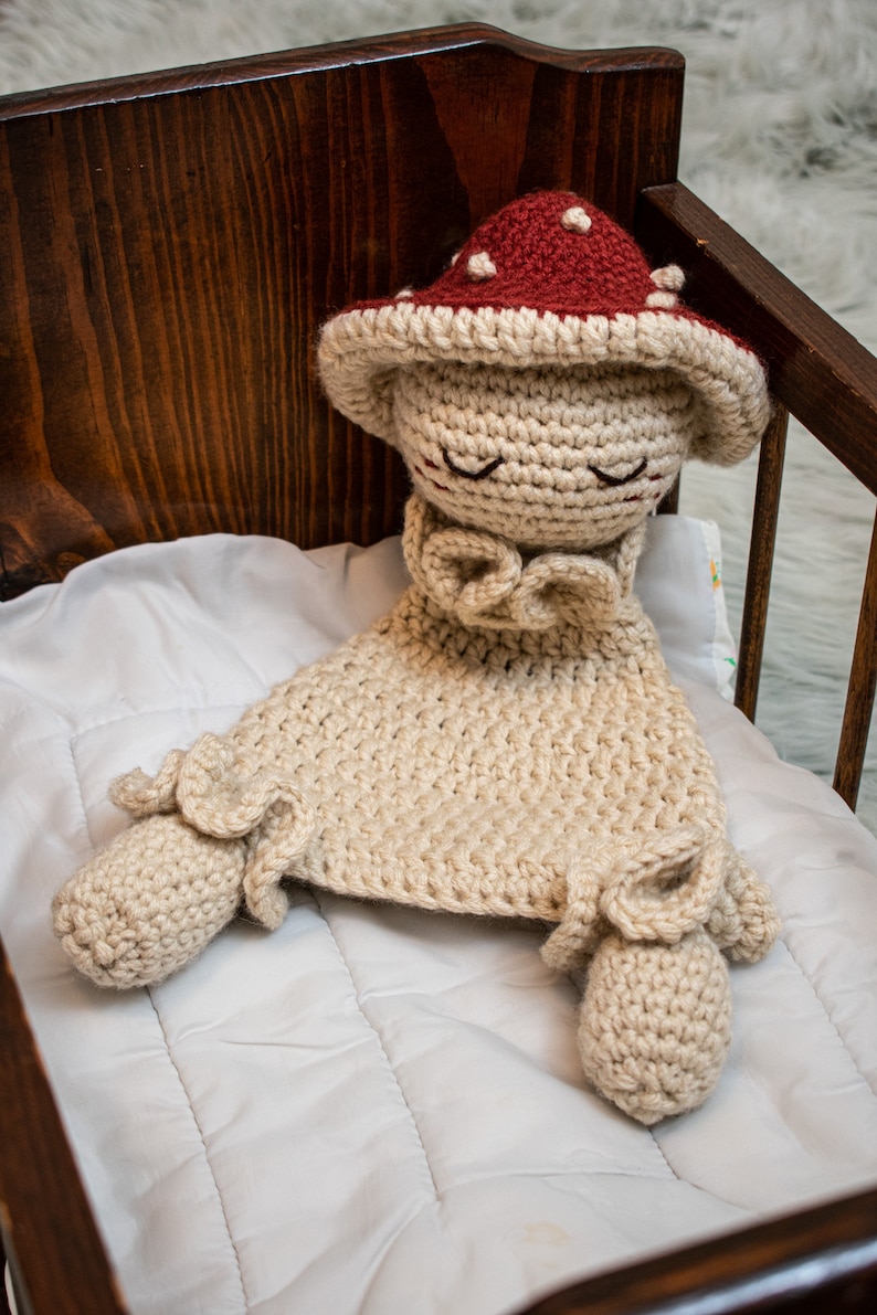 May include: A crocheted mushroom-shaped baby toy with a red and white hat, lying in a wooden crib with a white quilted mattress.
