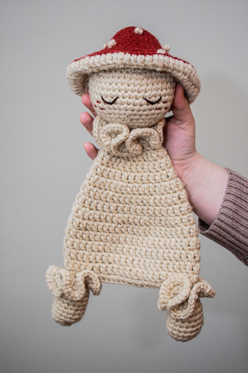 May include: A crocheted mushroom-shaped stuffed toy with a red cap and beige body. The toy has a sweet, smiling face with closed eyes and a ruffled collar.