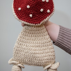 May include: A crocheted mushroom toy with a red cap and white spots. The mushroom has a beige stem and two small beige feet.