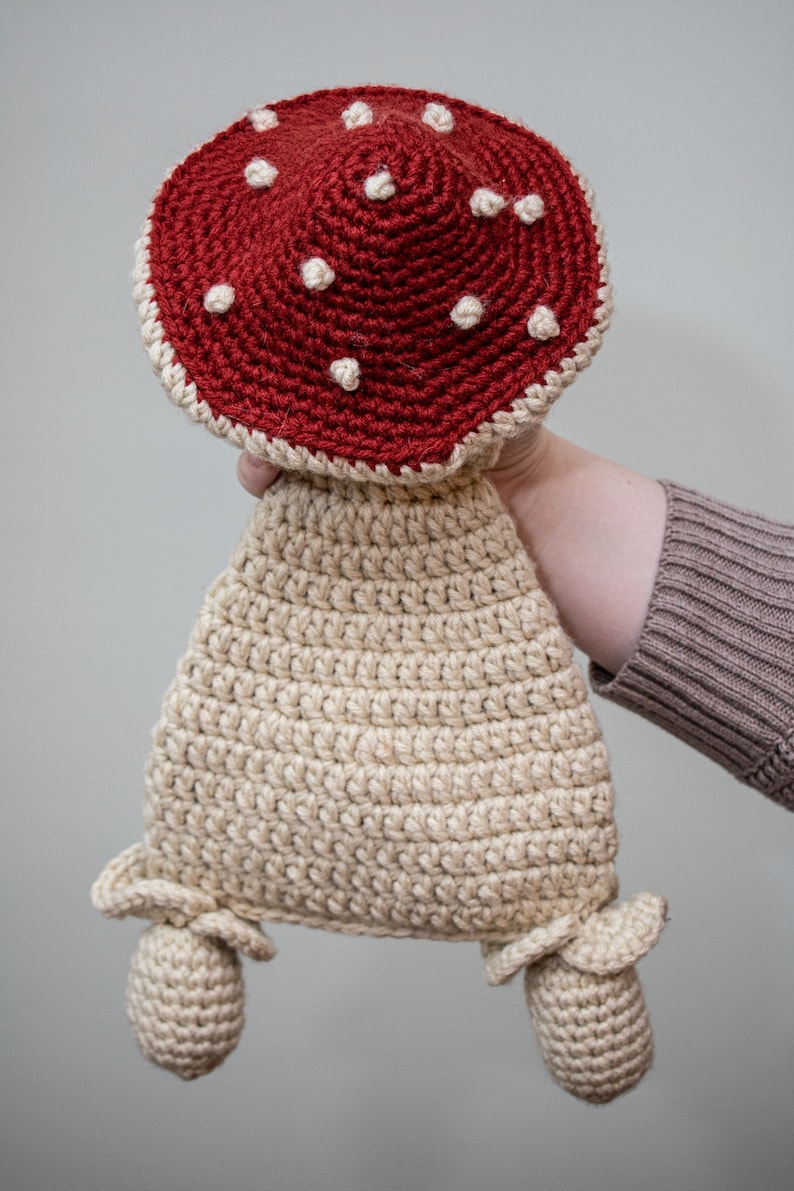 May include: A crocheted mushroom toy with a red cap and white spots. The mushroom has a beige stem and two small beige feet.