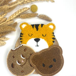 Könnte beinhalten: Drei Plüsch-Stickpatches: ein brauner Bär, ein Tiger mit schwarzen Streifen und ein brauner Keks mit einem Bissen. Die Patches befinden sich auf einer Holzoberfläche.