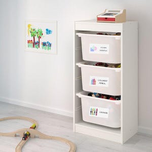 May include: White storage unit with three drawers labeled "Foam Shapes", "Colored Pencil", and "Crayon". The drawers are filled with colorful toys. A wooden train track is on the floor in front of the unit.