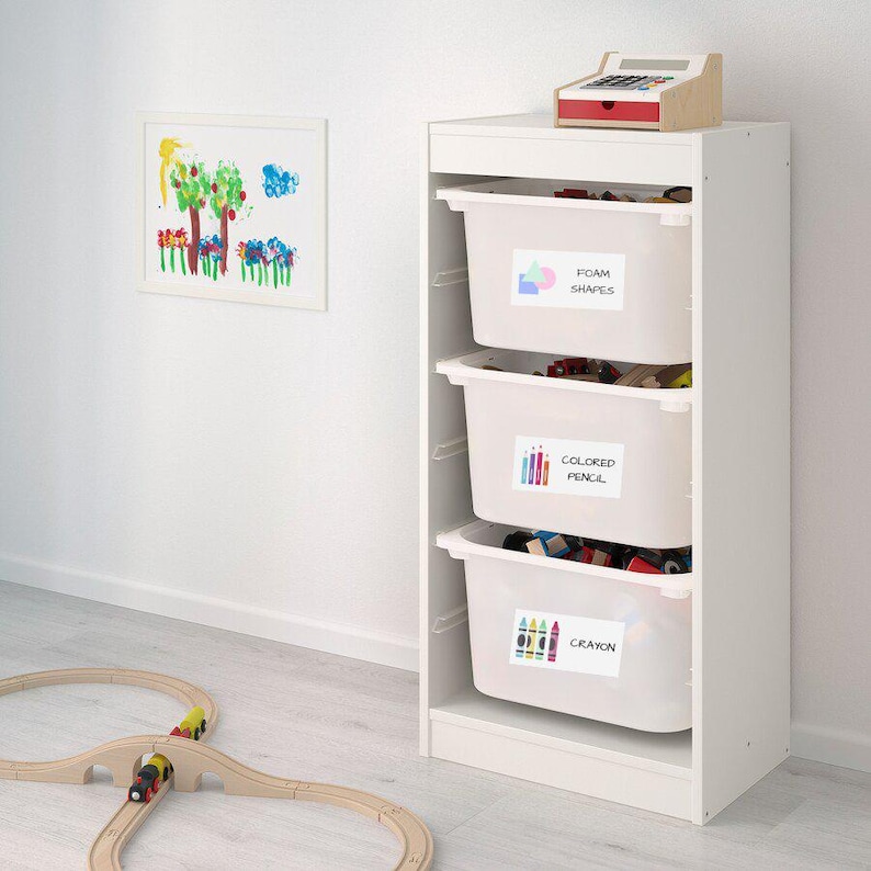 May include: White storage unit with three drawers labeled "Foam Shapes", "Colored Pencil", and "Crayon". The drawers are filled with colorful toys. A wooden train track is on the floor in front of the unit.