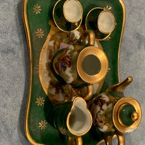 May include: A miniature porcelain tea set with a green and gold tray. The set includes a teapot, creamer, sugar bowl, and two cups and saucers. The porcelain is decorated with floral and landscape designs.
