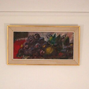 May include: An abstract painting of grapes and leaves in a gold frame. The artwork features a cluster of dark purple grapes, green leaves, and a yellow fruit against a red background. The painting is mounted on a beige mat.