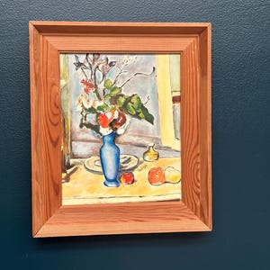 May include: A framed still life painting featuring a blue vase filled with flowers. Two small plates, two apples, and a pear are on a yellow table. The painting is in a light brown wooden frame.