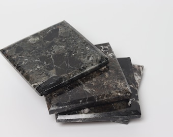 May include: Four square coasters made of dark gray marble with white veining. The coasters are stacked slightly, revealing their beveled edges. The marble has a polished finish, reflecting light against a white background.
