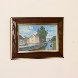 Puede incluir: Pintura al óleo de una escena de pueblo junto al agua en un marco de madera oscura. La obra representa un canal con pequeños botes, casas y árboles bajo un cielo azul con nubes. La pintura tiene una paleta de colores cálidos.