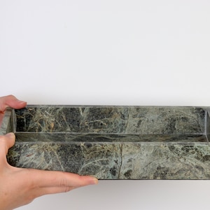 May include: A rectangular, dark green marble tray with a natural, veined pattern. The tray is held by two hands against a white background. The tray's dimensions are approximately 30 cm long, 10 cm wide, and 5 cm deep. Ideal for home decor.
