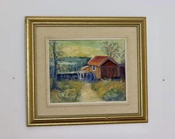 May include: Oil painting of a rural scene with a red-roofed building, a water wheel, and a path leading through a field. The artwork is framed in a gold-colored frame, adding a touch of elegance to the landscape depiction.