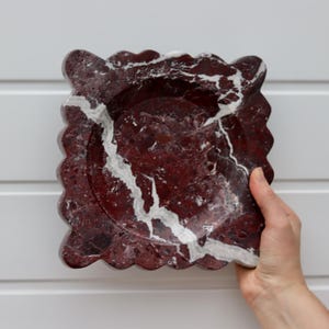 May include: A square, decorative dish with a scalloped edge. The dish is made of red marble with white veining. The dish is held by a hand against a white background.