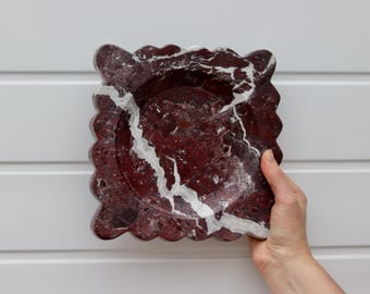 May include: A square, decorative dish with a scalloped edge. The dish is made of red marble with white veining. The dish is held by a hand against a white background.