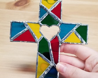 May include: A stained glass cross with a heart cut out in the center. The cross is made of red, yellow, blue, and green glass.