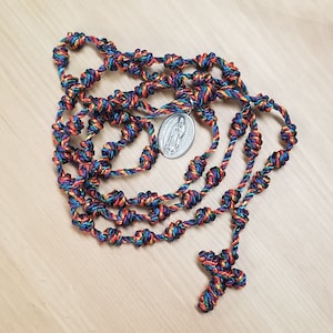 K&ouml;nnte beinhalten: Ein farbenfroher geknoteter Rosenkranz mit einem silbernen Anh&auml;nger, der das Bild der Jungfrau Maria zeigt. Der Rosenkranz besteht aus mehreren Str&auml;ngen geflochtener Schnur in den Farben Blau, Gr&uuml;n, Orange und Lila.