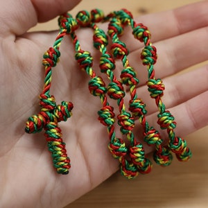 K&ouml;nnte beinhalten: Ein geknotetes Rosenkranzband aus gr&uuml;nem, rotem und gelbem geflochtenem Seil. Der Rosenkranz hat ein gekreuztes Ende.