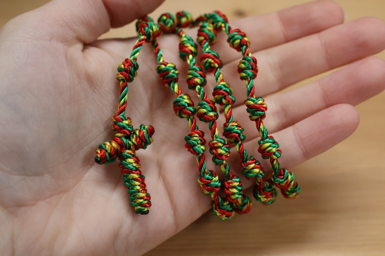K&ouml;nnte beinhalten: Ein geknotetes Rosenkranzband aus gr&uuml;nem, rotem und gelbem geflochtenem Seil. Der Rosenkranz hat ein gekreuztes Ende.