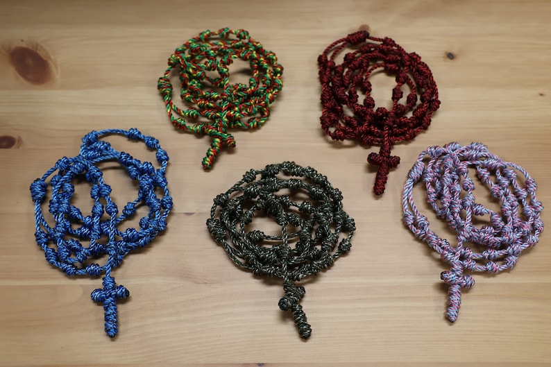 K&ouml;nnte beinhalten: F&uuml;nf gekn&uuml;pfte Rosenkr&auml;nze aus Seil mit Kreuzen. Die Rosenkr&auml;nze sind in verschiedenen Farben: blau, gr&uuml;n, rot und lila.