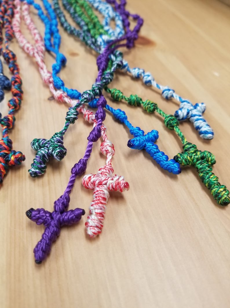 K&ouml;nnte beinhalten: Eine Sammlung bunter, gekn&uuml;pfter Rosenkr&auml;nze aus Seil mit verschiedenen farbigen Kreuzen. Die Rosenkr&auml;nze bestehen aus verschiedenen Farben von Schnur, darunter Lila, Blau, Gr&uuml;n, Rot und Wei&szlig;.
