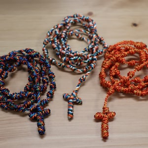 K&ouml;nnte beinhalten: Drei gekn&uuml;pfte Rosenkr&auml;nze aus Seil mit Kreuzen. Einer ist blau, wei&szlig; und orange. Einer ist orange und gold. Einer ist wei&szlig; und gold.