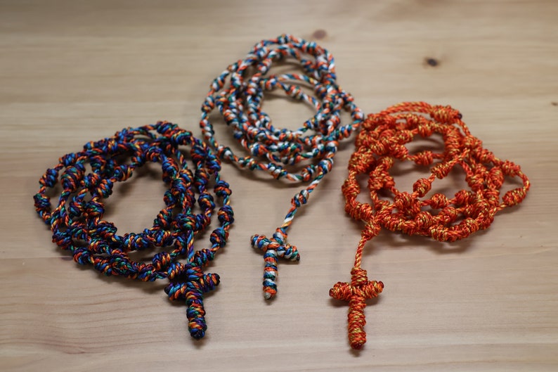 K&ouml;nnte beinhalten: Drei gekn&uuml;pfte Rosenkr&auml;nze aus Seil mit Kreuzen. Einer ist blau, wei&szlig; und orange. Einer ist orange und gold. Einer ist wei&szlig; und gold.