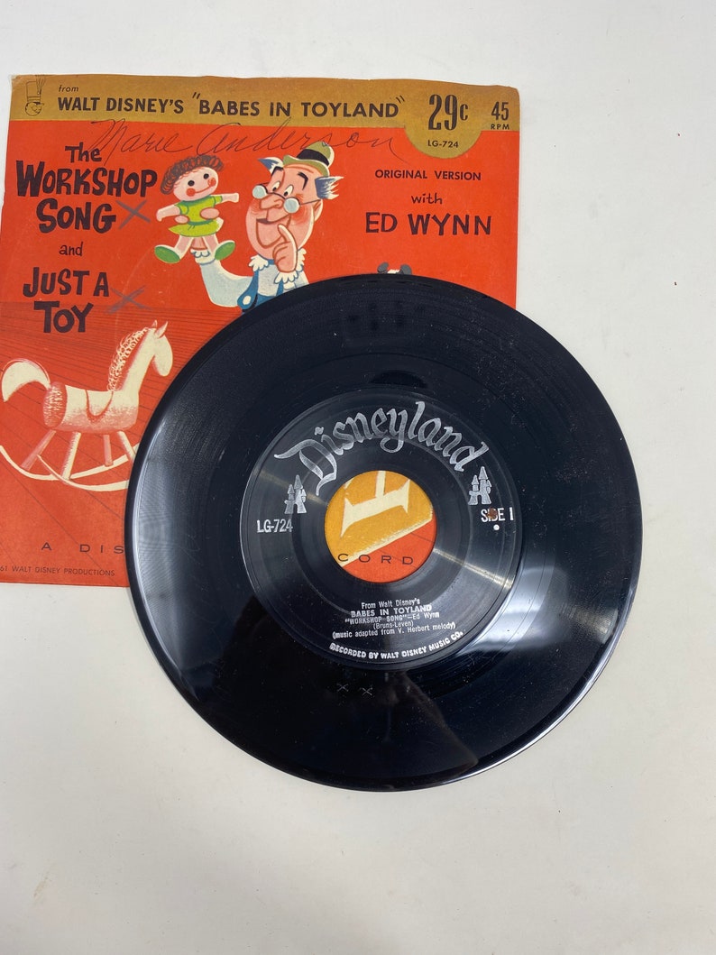 K&ouml;nnte beinhalten: Eine schwarze Schallplatte mit einem roten und gelben Etikett, auf dem "Disneyland" und "Seite 1" steht. Die Schallplatte ist aus Walt Disneys "Babes in Toyland" und enth&auml;lt den Song "The Workshop Song" mit Ed Wynn. Die Schallplatte ist 45 U/min und stammt von Walt Disney Productions.