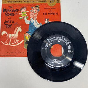 K&ouml;nnte beinhalten: Eine Vintage-Schallplatte mit 45 U/min mit einer schwarzen Vinyl-Scheibe und einer roten, wei&szlig;en und blauen Papierh&uuml;lle. Die H&uuml;lle zeigt eine Comic-Illustration eines Spielzeugmachers und eines Schaukelpferdes. Die Schallplatte tr&auml;gt den Titel "The Workshop Song and Just a Toy" aus Walt Disneys "Babes in Toyland".