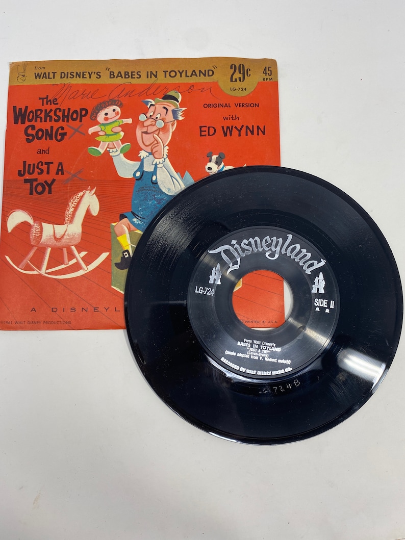K&ouml;nnte beinhalten: Eine Vintage-Schallplatte mit 45 U/min mit einer schwarzen Vinyl-Scheibe und einer roten, wei&szlig;en und blauen Papierh&uuml;lle. Die H&uuml;lle zeigt eine Comic-Illustration eines Spielzeugmachers und eines Schaukelpferdes. Die Schallplatte tr&auml;gt den Titel "The Workshop Song and Just a Toy" aus Walt Disneys "Babes in Toyland".
