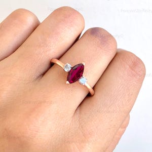 May include: A rose gold ring featuring a marquise-cut, deep red gemstone as the focal point, flanked by two smaller, round, clear gemstones. The ring's band is slender and elegant, showcasing a minimalist design.