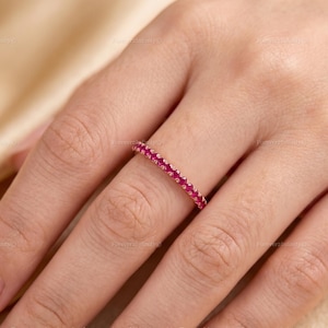 May include: A thin, delicate ring featuring a continuous band of small, round, vibrant pinkish-red gemstones set in a polished metal frame. The band is uniform in width and showcases a series of closely spaced, faceted stones that catch the light.