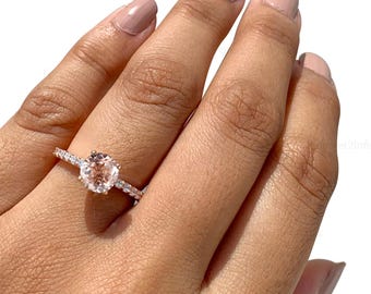 May include: Rose gold ring featuring a round, peach-colored gemstone as the centerpiece. The band is adorned with smaller, clear gemstones. The ring is set on a hand with a neutral nail polish.