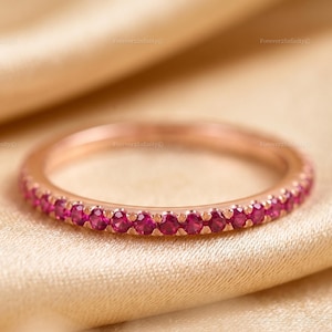 May include: A rose gold eternity band featuring a continuous row of small, round, vibrant pinkish-red gemstones set along the outer circumference of the metal ring. The band has a polished finish and a slim, uniform profile throughout its design.