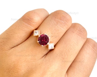 May include: A gold ring with a large, round, dark red gemstone surrounded by smaller, clear gemstones. The ring is on a person's finger.
