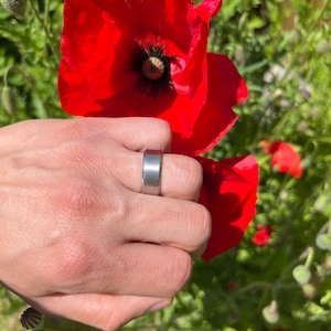 Puede incluir: Un anillo de banda de plata con acabado cepillado. El anillo se lleva en el dedo índice de una mano que sostiene una flor de amapola roja.