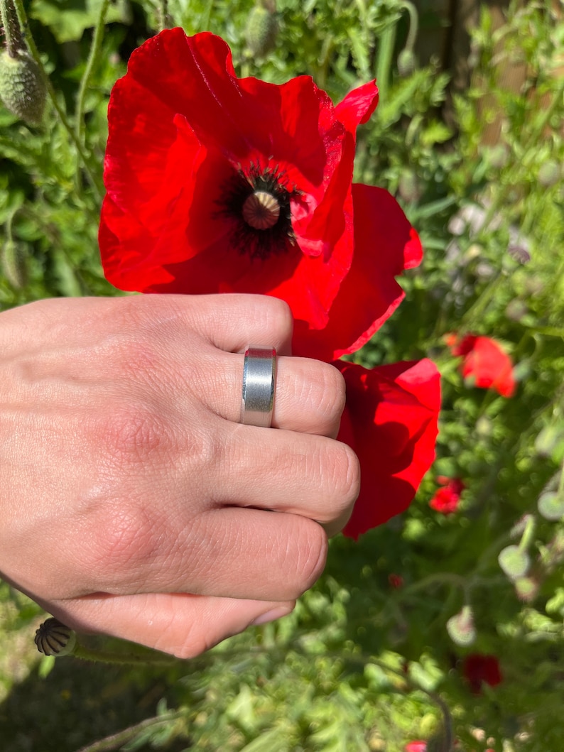 May include: A silver band ring with a brushed finish. The ring is worn on the index finger of a hand holding a red poppy flower.