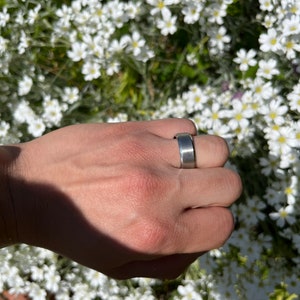May include: A silver band ring on a hand with a white flower background.