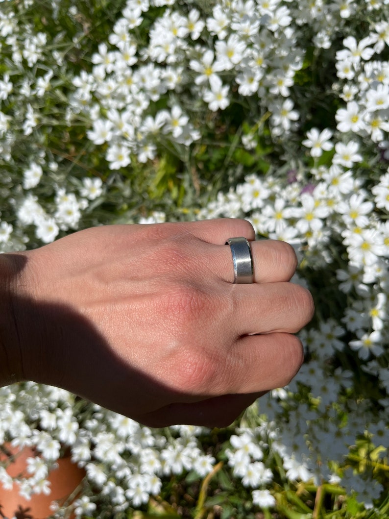 May include: A silver band ring on a hand with a white flower background.